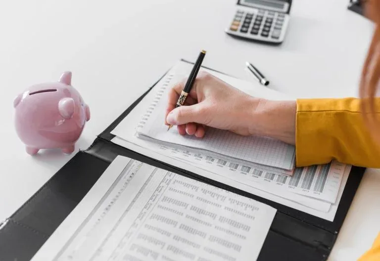 Main écrivant près d’une tirelire et d’une calculatrice.